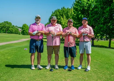 Four men wearing pink golf polos smile while holding their clubs